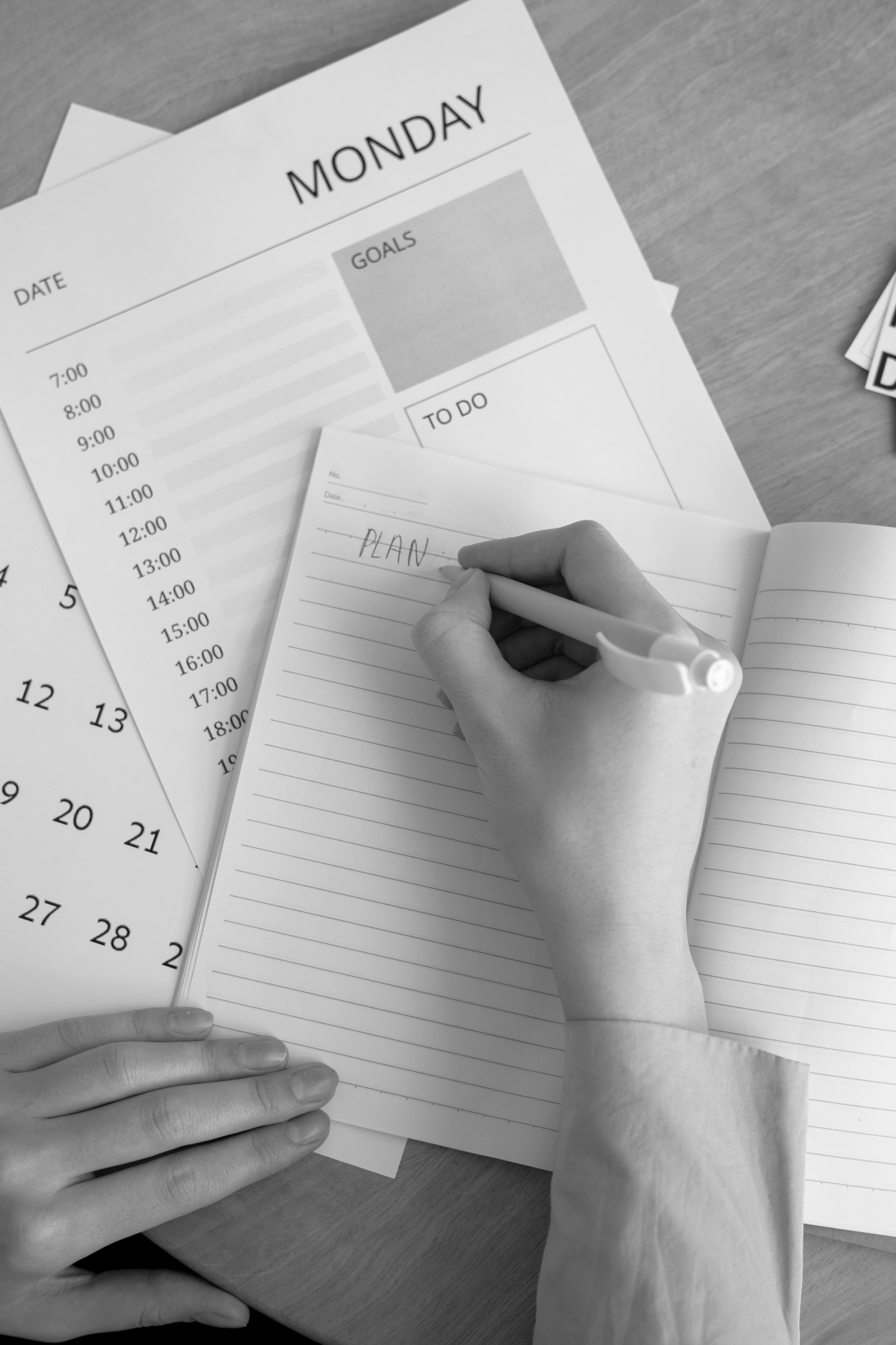 A hand writing 'PLAN' in a notebook over a Monday schedule and a calendar.